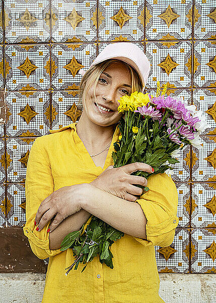 Glückliche junge Frau mit Mütze und Blumenstrauß vor gemusterter Wand