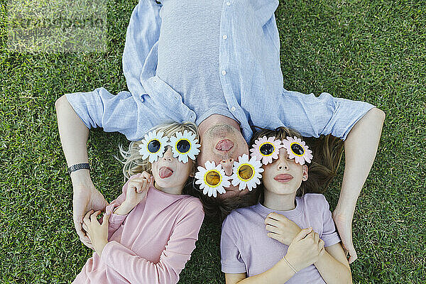 Verspielter Vater und Töchter mit Blumensonnenbrille entspannen sich und haben Spaß im Gras