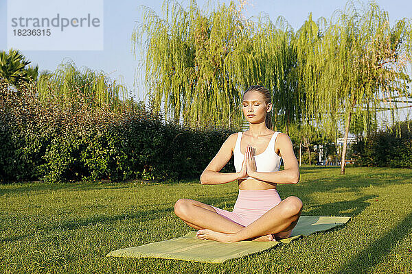 Frau mit geschlossenen Augen übt Padmasana-Pose auf Gymnastikmatte im Park