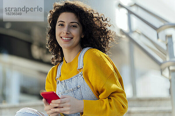 Lächelnde Frau mit lockigem Haar  die ihr Mobiltelefon in der Hand hält