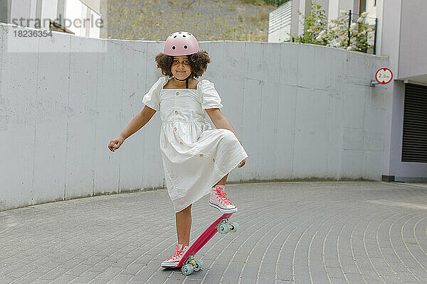 Lächelndes Mädchen mit Helm  das auf dem Fußweg Skateboard fährt