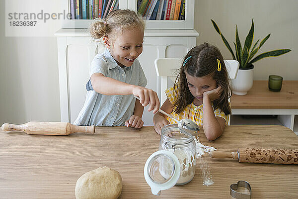 Happy girl pouring flour on table with friend at home
