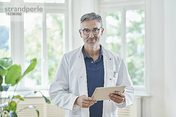 Lächelnder Arzt steht mit Tablet-PC in der Arztpraxis