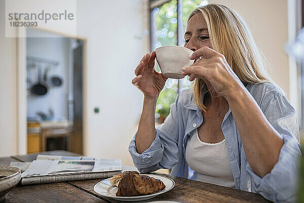 Reife Frau trinkt Kaffee mit Croissant auf dem Tisch zu Hause