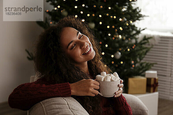 Lächelnde junge Frau hält eine Tasse Marshmallow-Kakao in der Hand und sitzt zu Hause auf dem Sofa