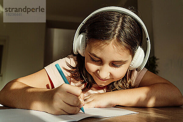 Lächelndes Mädchen mit kabellosen Kopfhörern  das am Schreibtisch in ein Buch schreibt