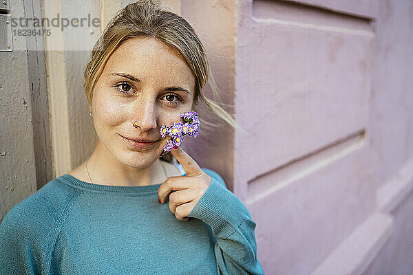 Lächelnde junge Frau hält kleine Blumen im Gesicht vor der Wand