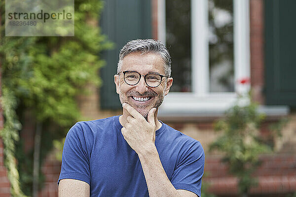 Lächelnder Mann mit Brille und Hand am Kinn vor dem Haus