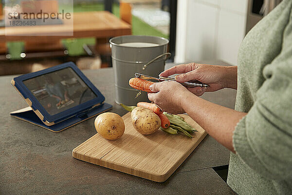 Frau mit Tablet-PC schält Karotten und bereitet zu Hause eine Mahlzeit zu
