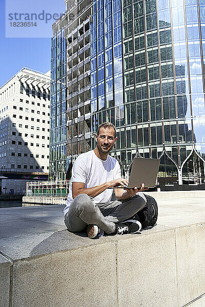 Lächelnder Mann mit Laptop sitzt vor Gebäuden