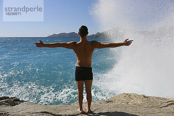Mann mit ausgestreckten Armen blickt auf Wellen  die auf Felsen schlagen