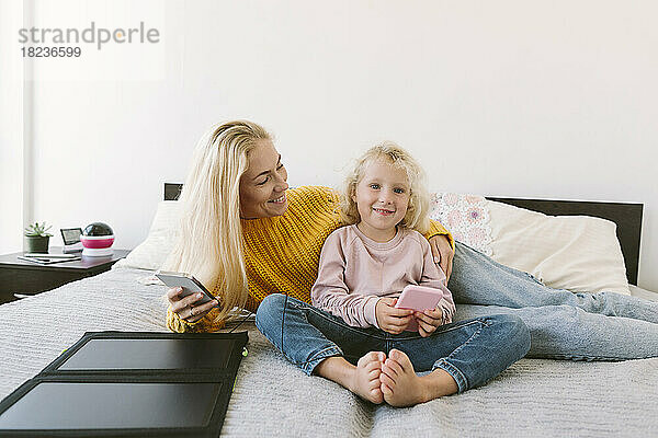 Lächelnde Mutter und Tochter sitzen mit Smartphones auf dem Bett im Schlafzimmer