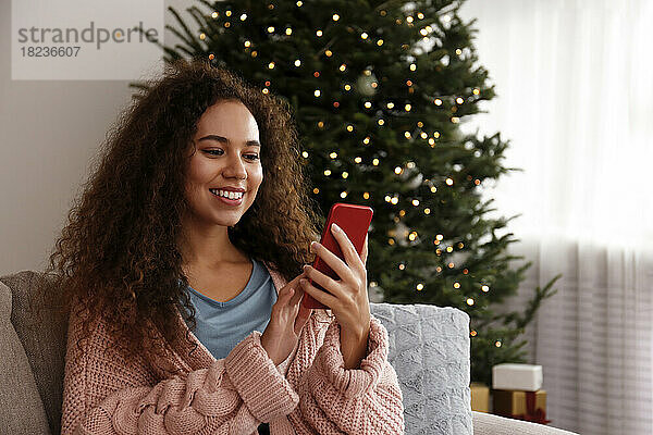 Glückliche junge Frau benutzt Smartphone vor dem Weihnachtsbaum zu Hause