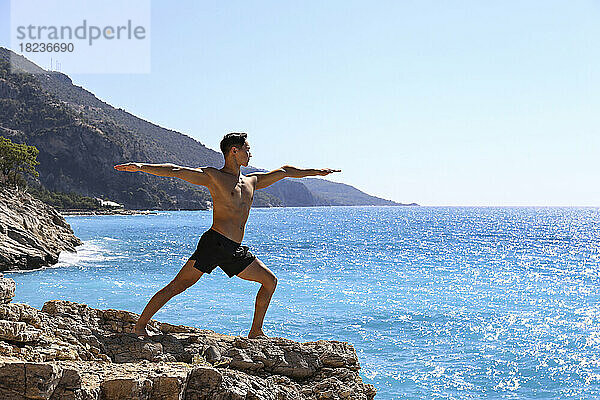 Shirtless man practicing Virabhadrasana yoga pose standing on rock near sea