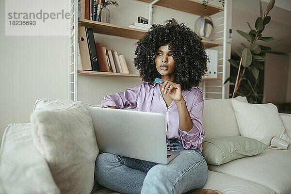 Nachdenkliche Frau mit Laptop und Kreditkarte sitzt im Wohnzimmer