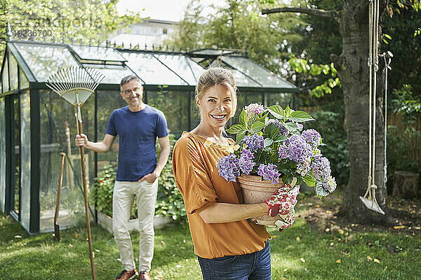 Lächelnde Frau hält Topfpflanze mit Mann im Hintergrund im Hinterhof