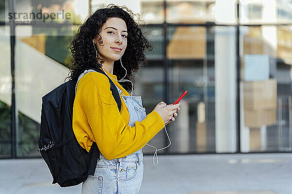 Schöne Frau mit Rucksack hält ihr Mobiltelefon in der Hand und hört auf dem Fußweg Musik über In-Ear-Kopfhörer