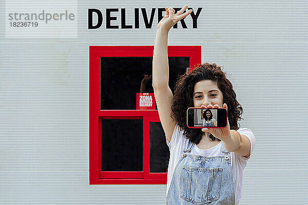 Frau macht Selfie mit Handy vor Wand mit Lieferschild