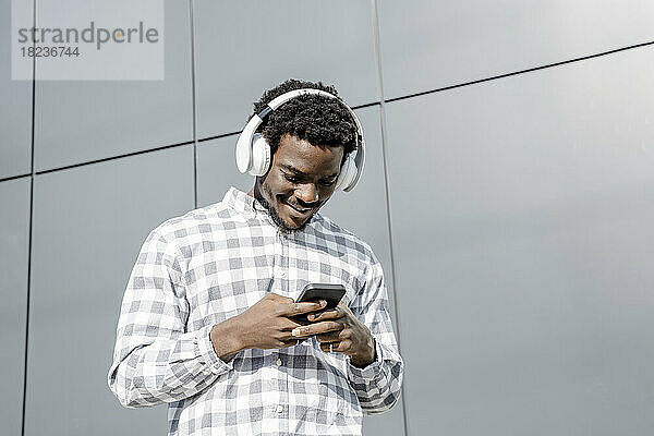 Lächelnder Mann mit Kopfhörern und Smartphone an der Wand