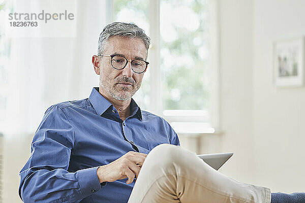Reifer Geschäftsmann schaut zu Hause im Sessel auf den Tablet-PC