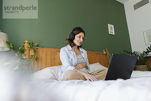 Lächelnde schwangere Geschäftsfrau mit kabellosen Kopfhörern und Laptop auf dem Bett im Schlafzimmer