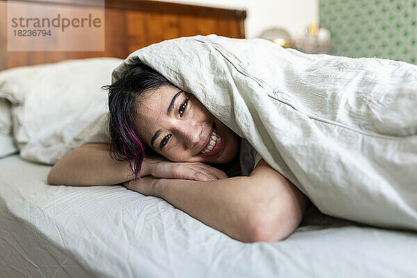 Glückliche junge Frau  die zu Hause mit Decke auf dem Bett liegt