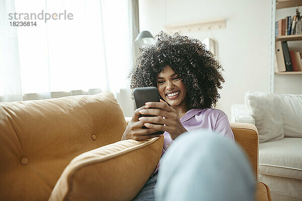 Lächelnde Frau benutzt Smartphone auf Sofa im Wohnzimmer