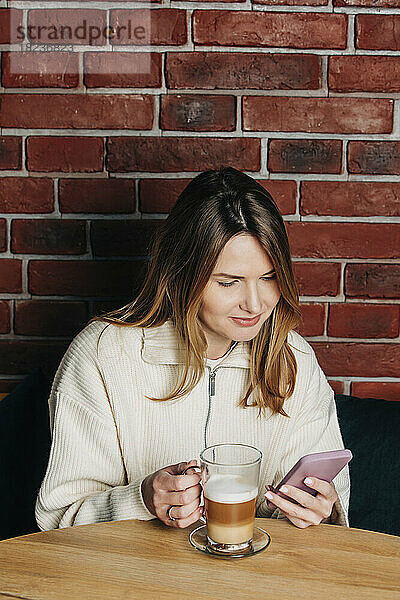 Frau benutzt Mobiltelefon und trinkt Cappuccino am Tisch im Café