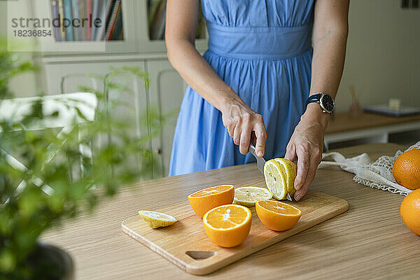 Hände einer Frau  die zu Hause Zitronen und Orangen auf dem Tisch schneidet