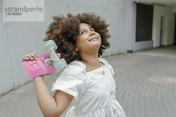 Lächelndes Mädchen mit Afro-Frisur lehnt Kopf auf Skateboard am Fußweg