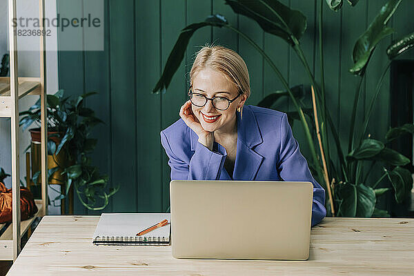 Lächelnde Geschäftsfrau mit Laptop sitzt am Schreibtisch im Heimbüro