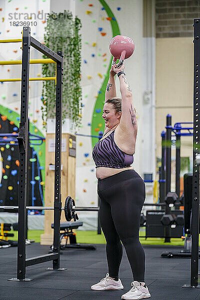 Lächelnde übergewichtige Frau mit erhobenen Armen beim Kettlebellheben im Fitnessstudio