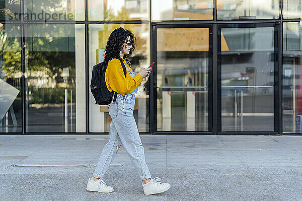 Frau mit Rucksack benutzt Smartphone vor Glasgebäude auf Fußweg