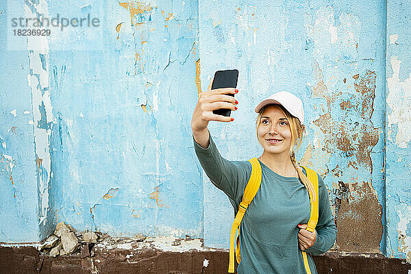 Lächelnde Frau mit Rucksack macht Selfie mit Smartphone vor der Wand