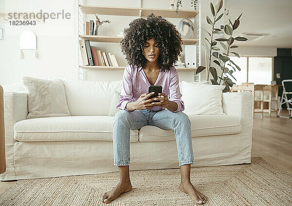 Junge Frau benutzt Mobiltelefon und sitzt auf dem Sofa im Wohnzimmer