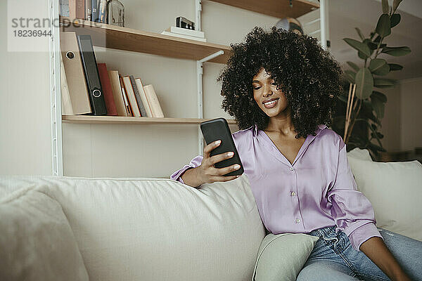 Junge Frau mit Afro-Frisur benutzt Mobiltelefon im Wohnzimmer