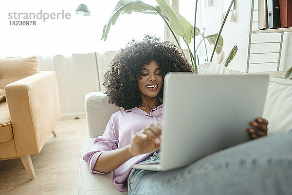Lächelnde Afro-Frau mit Laptop  die zu Hause auf dem Sofa liegt