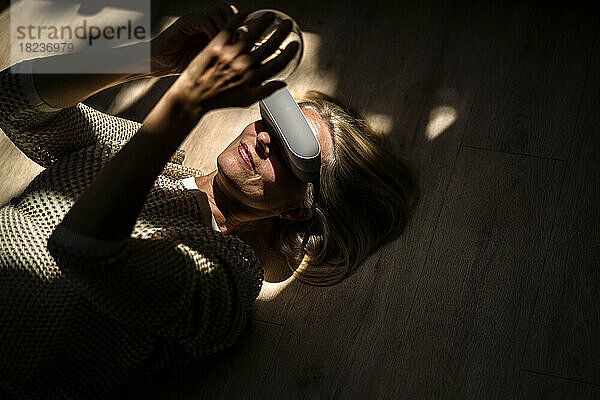 Reife Frau mit VR-Brille und Glühbirne in der Hand liegt zu Hause