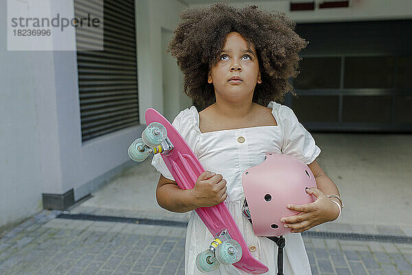 Nachdenkliches Mädchen mit Skateboard und Helm steht vor dem Eingang