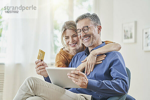 Lächelnde Frau umarmt Mann mit Kreditkarte beim Online-Shopping per Tablet-PC zu Hause