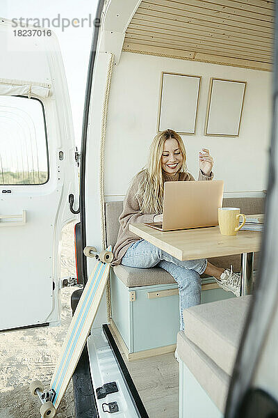 Lächelnder junger digitaler Nomade  der im Wohnmobil am Laptop arbeitet