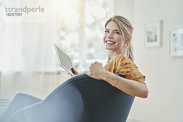 Glückliche Frau hält Tablet-PC im Sessel zu Hause