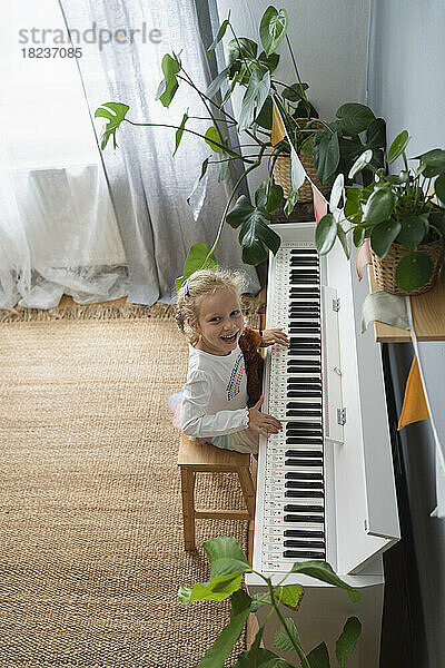 Glückliches Mädchen  das zu Hause inmitten von Pflanzen Klavier spielt