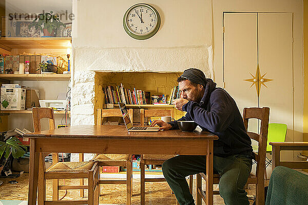 Freiberufler isst Mahlzeit mit Laptop am Schreibtisch zu Hause