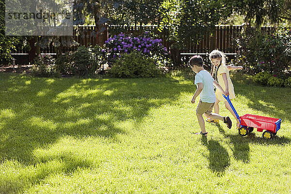 Playful girl pulling toy wagon with brother on sunny day in back yard