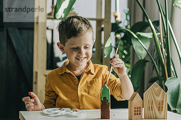 Fröhlicher Junge malt Holzmodellhäuser am Tisch