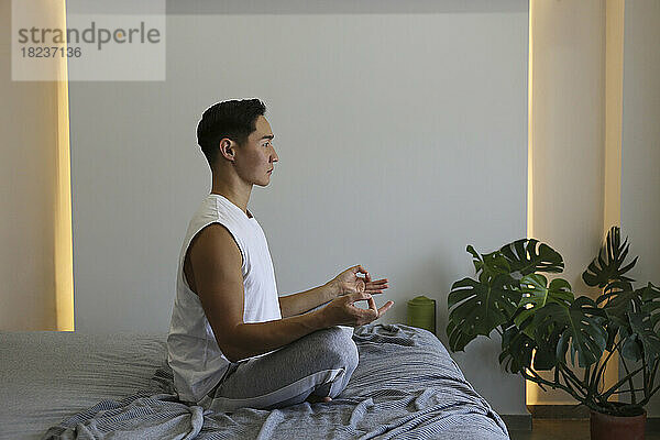 Junger Mann praktiziert Yoga und sitzt zu Hause im Bett