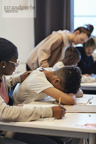 Teenager-Mädchen  das einen deprimierten männlichen Schüler tröstet  der seinen Kopf im Klassenzimmer auf den Schreibtisch legt