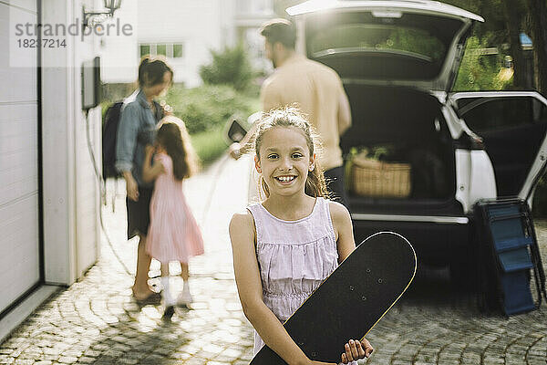 Lächelndes Mädchen hält Skateboard mit Familie im Hintergrund