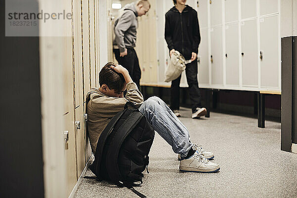 Depressiver Junge in voller Länge mit Rucksack im Schulkorridor sitzend